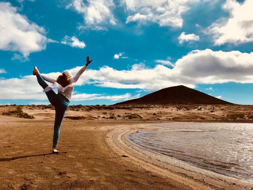 A woman practices yoga by a desert lake with a mountain backdrop under a sunny blue sky.